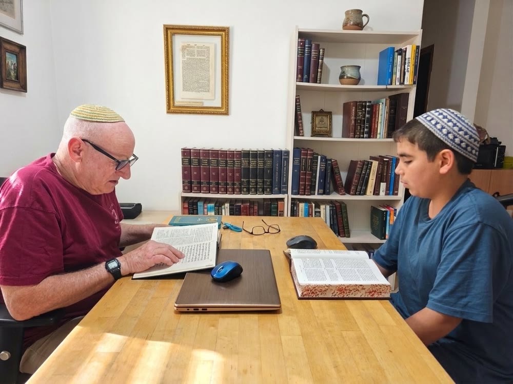 סבא ונכד לומדים תנ"ך יחד ליד שולחן, ומאחוריהם תלוי על הקיר דף בומברג ממוסגר